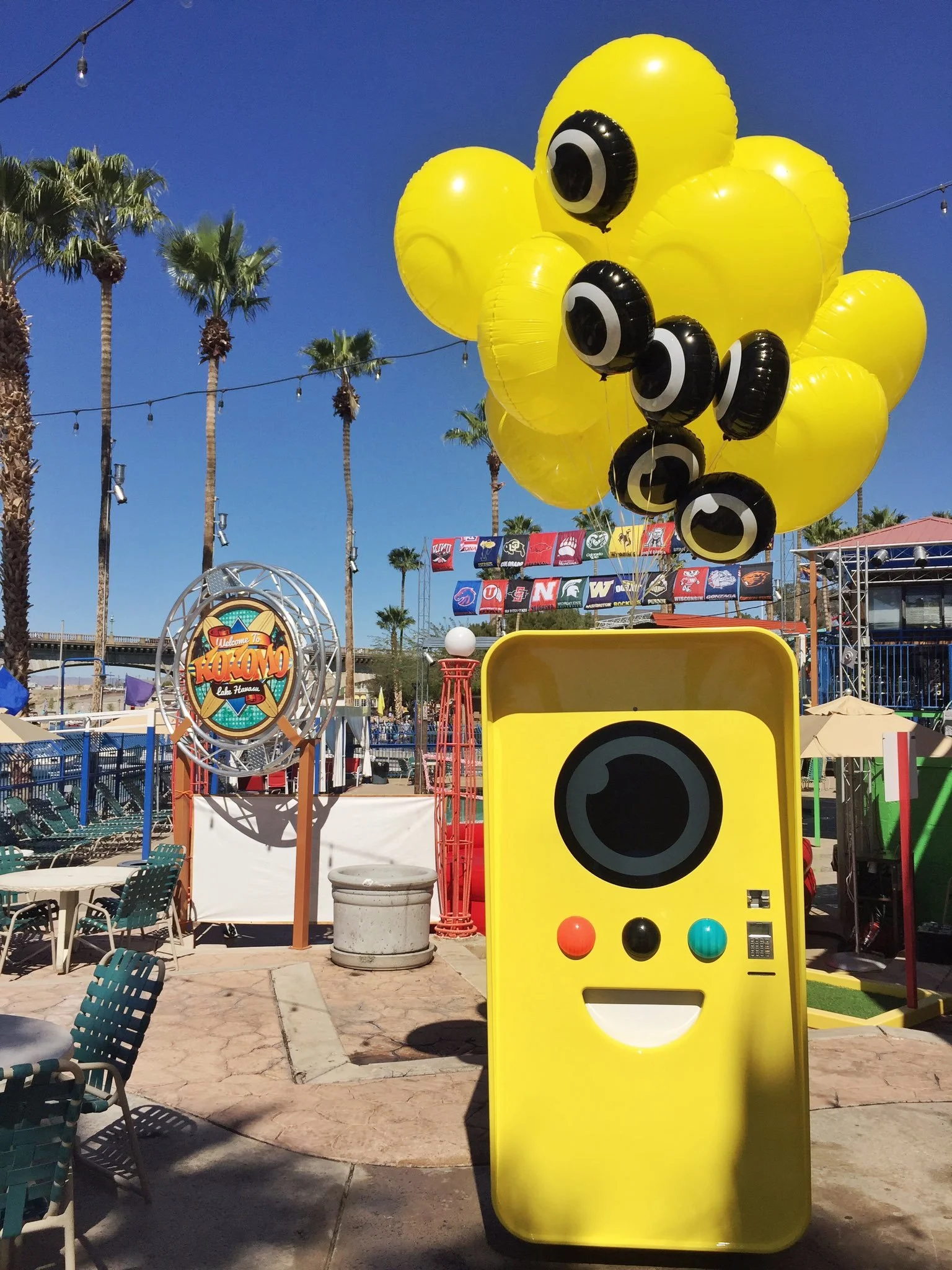 Yellow Snapbot vending machine with eyeball balloons at a palm-lined poolside venue under blue sky